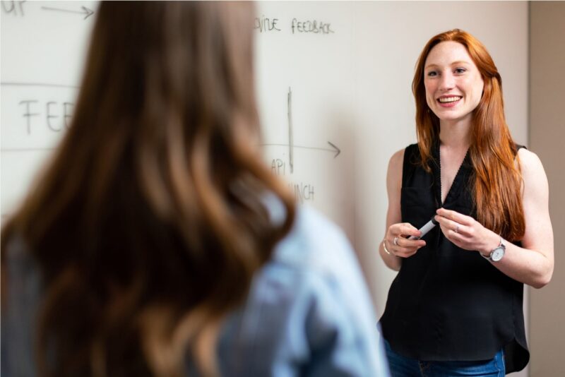 A female teacher smiles while standing in front of a whiteboard, shown over the shoulder of a student