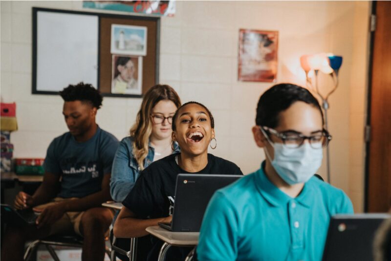 A girl using a laptop in class laughs, with a masked student in front of her